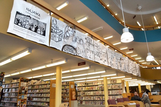 Photo taken at the installation of "Najawa: A Story of Palestine" in Burlington, VT's Fletcher Free Library. The full comic is in view, hanging from a second-story banister.