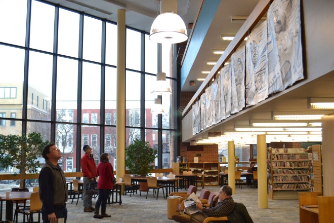 Photo taken at the opening of "Najawa: A Story of Palestine" in Burlington, VT's Fletcher Free Library. A group of people are looking up from the main room to where the comic hangs from a second-story banister.