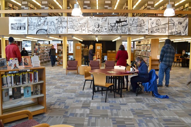 Photo taken at the installation of "Najawa: A Story of Palestine" in Burlington, VT's Fletcher Free Library. A group of people are looking up from the main room to where the comic hangs from a second-story banister.