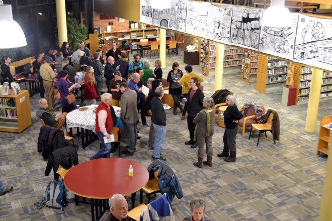 Photo taken at the opening of "Najawa: A Story of Palestine" in Burlington, VT's Fletcher Free Library. A group of people are gathered in the main room, where the comic hangs from a second-story banister.