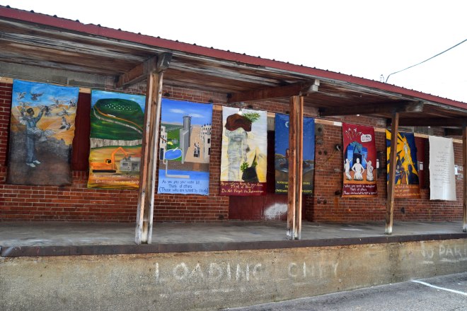 Photo of the "Think of Others" exhibit at Art Hop. 8 of the canvas panels are shown hanging on a loading dock beneath an overhang.