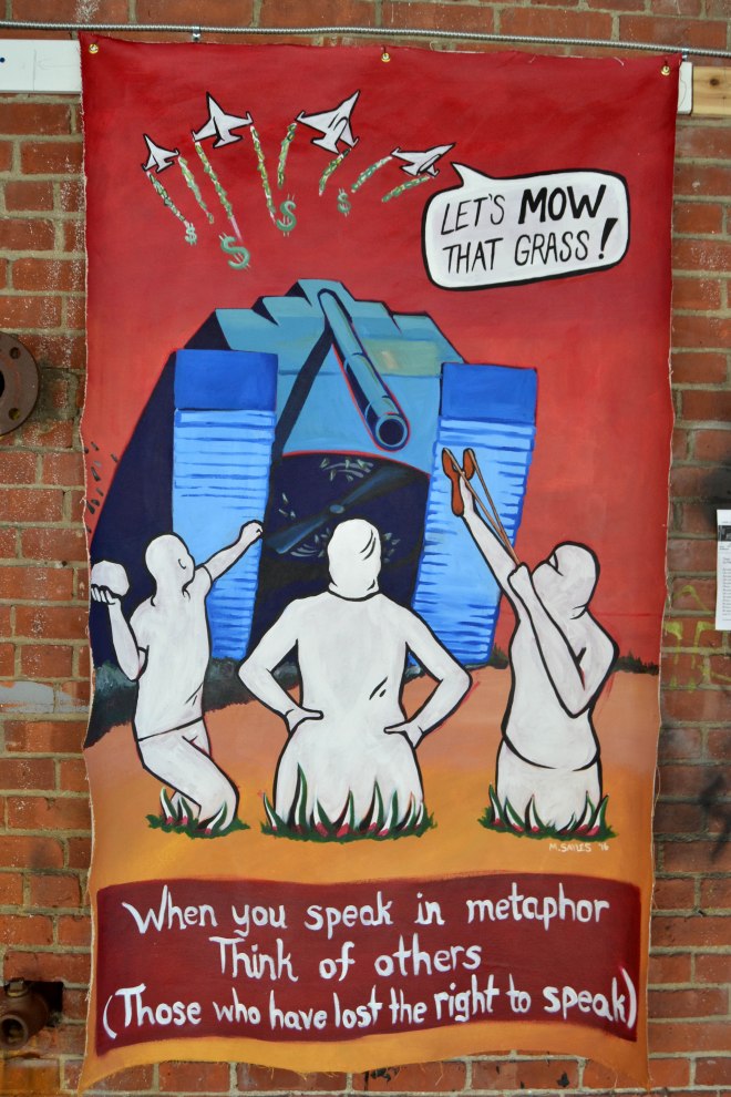 Photo of a panel from the Think of Others exhibit. Shows three Palestinians planted in the ground, with red, white, and green grass growing around them, in the colors of the Palestinian flag. A larger-than-life tank towers above them, pointing down at them. Fighter jets burning cash fly by, and one pilot calls out "Let's Mow that grass!" Reads: "When you speak in metaphor, think of others (Those who have lost the right to speak)."