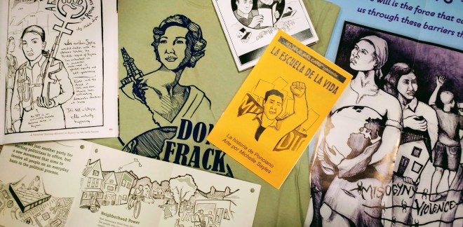 Photograph of artwork samples by Michelle Sayles. Includes a t-shirt printed with the "Don't Frack Your Mother" design; a poster print; Spanish-language zine of "The School of Life" comic; and a Rojava comic.